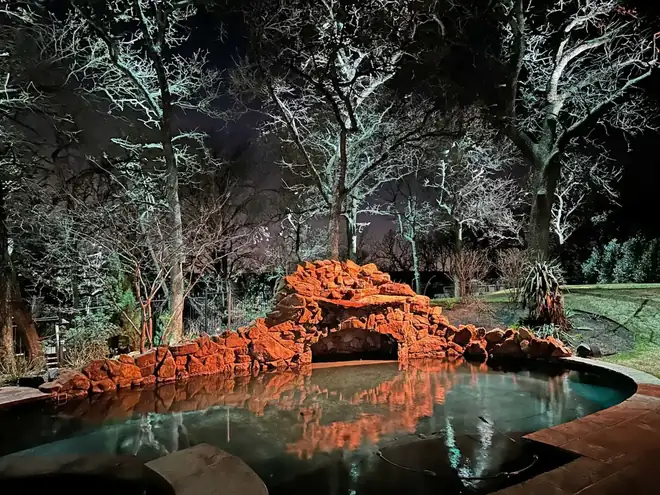 Stone waterfall grotto lit warm orange above a still pool, framed by moonlit bare oaks in Corinth, Texas