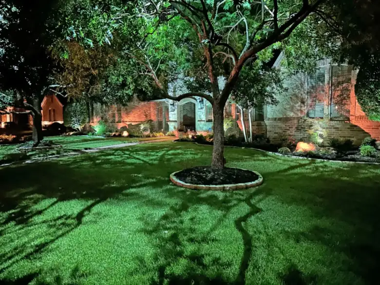 Moonlighting downlights casting tree-branch shadows across a front lawn in Preston Hollow, Dallas, with the brick facade washed warm behind